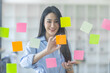© David - Smiling business Asian people thinking and use sticky notes on glass wall in office with her colleague in background.