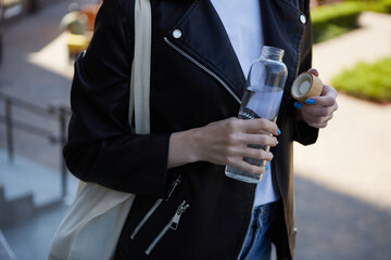 Wall Mural - Thirsty young woman drinking mineral water from a glass bottle. Responsible female person using a reusable water bottle and wearing a cotton tote bag on shoulder