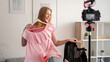 © golubovy - Showing clothes. Filming video. Fashion advice. Cheerful playful young woman trying pink dress on hanger recording vlog on camera on tripod in white room.