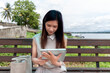 © 2D_Jungle - Asian woman sitting on a scenic bench by the river along with playing the tablet with intention