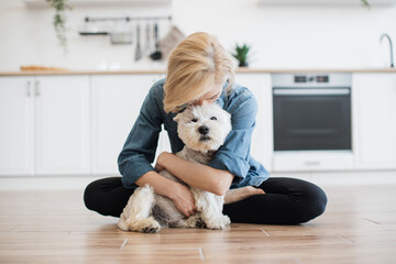 Wall Mural - Portrait of smiling woman hugging gently adorable dog while posing on wooden floor with legs crossed. Happy female owner showing care for beloved pet before going for after dinner walk to park.
