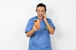 © Roquillo - Happy Young doctor woman wearing blue uniform over isolated background feels good while focused in screen of smartphone. People, technology, lifestyle