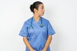 © Roquillo - Close up side profile photo Young doctor woman wearing blue uniform over isolated background not smiling attentive listen concentrated