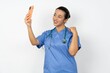 © Roquillo - Young doctor woman wearing blue uniform over isolated background smiling and taking a selfie ready to post it on her social media.