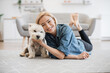 Full length portrait of lovely smiling woman in denim shirt caressing adorable little Westie on kitchen floor of apartment. Cheerful pet keeper posing with good-natured small animal in home interior.