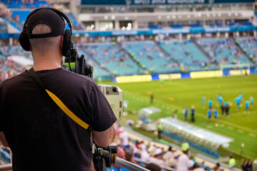 Professional cameraman in a black T-shirt with a camera shoots a football match in the stadium. Cameraman with professional sound recording equipment shoots a report