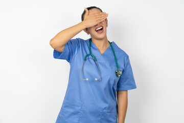 Wall Mural - Beautiful doctor woman standing over white studio background smiling and laughing with hand on face covering eyes for surprise. Blind concept.