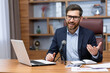© Liubomir - Portrait of successful man inside office, businessman with professional microphone recording online training course and audio podcasts, using laptop sitting at desk working, looking at camera.