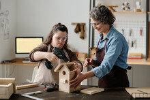 Girl With Down Syndrome Using Drill To Make Birdhouse Together With Master In Workshop