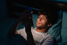 Insomnia, Addiction Concept. Sleepy Exhausted Man Lying In Bed Using Smartphone, Can Not Sleep. Handsome Guy In Bed Scrolling Through Social Networks On Mobile Phone At Night In Dark