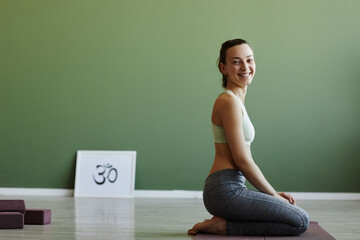 Wall Mural - Side view of fit young woman doing yoga indoors and smiling at camera happily with copy space