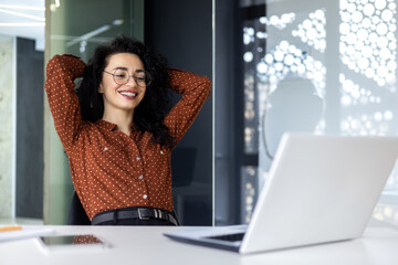 Happy and smiling successful hispanic business woman finished work satisfied with work result and achievement, hands behind head and smiling close up