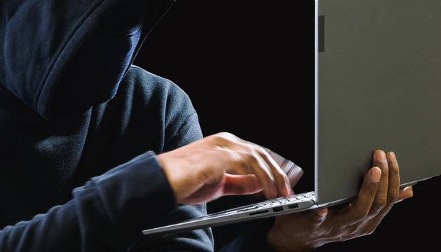 Hacker spy man one person in black hoodie sitting on a table looking computer laptop used login password attack security to circulate data digital in internet network system, night dark background.