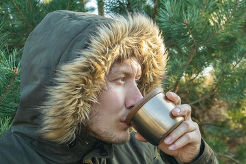 A young man in a hood is drinking hot tea from a mug. Tourist trip. Outdoor recreation