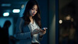 © Mauricio - Young serious busy Asian business woman executive using mobile smartphone at night in dark office. Professional businesswoman holding smartphone, working on cellphone. Generative AI