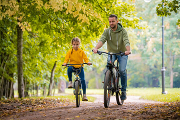 Happy father and daughter ride bicycles in autumn park on sunny day.