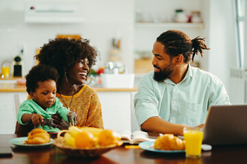 A cheerful african american family sitting at the breakfast table and talking while a husband working on a laptop.