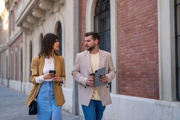 Serious businessman and businesswoman discussing new business strategy project plan while walking outside on the way to meeting at work, looking at each other holding mobile phone, tablet and coffee.