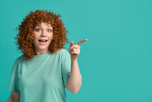 Surprised Excited Attractive Young Ginger Woman Wearing Casual Clothes And Smiling Pointing Finger At Turquoise Copy Space Turning Attention On Something New Looking At Camera Isolated Over Background