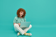 Smiling Cheerful Smart Young Ginger Woman Wearing Casual Teenage Clothes Using Laptop Computer Sitting On Floor In Lotus Pose Looking At Camera Isolated Over Turquoise Background With Copy Space