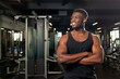 © Богдан Маліцький - young African American man stands with crossed arms in dark gym and smiles, fitness trainer stands in fitness club