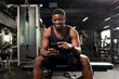 © Богдан Маліцький - young athletic african american man sits in dark gym and uses smartphone, sports guy types online on the phone