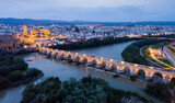 Scenic aerial view of lighted Roman bridge across Guadalquivir river and Mezquita-Catedral on background with Cordoba cityscape at twilight, Spain..