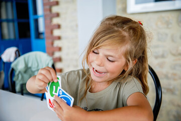 Wall Mural - Excited smiling cute preschool girl playing card game. Happy healthy child plays with family. Creative indoors leisure and education of kid. Family activity at home.