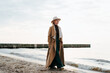 © Sergio - Thoughtful stylish mature woman walking on beach by sea. Alone senior woman in brown coat on seashore outdoors