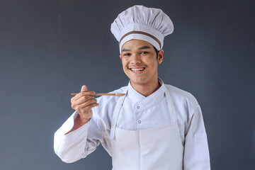 Young Asian male chef in white toque and apron with wooden spoon tasting the food isolated over grey background.