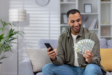 Happy Hispanic Man Holding Cash Dollars And Mobile Phone. Sitting At Home On The Sofa And Admiringly Looking At The Fan Of Money