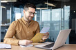 © Tetiana - A young man businessman sits smiling in the office at the table and holds in his hands and reads documents, a contract, a successful deal that he received by mail