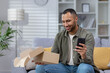 © Tetiana - A happy young Latin American man is sitting on a sofa, holding a phone in his hand and is happy with a received gift, a parcel that was delivered to his home