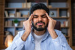 © Ivan - Tired young man indian businessman feels severe headache migraine. He sits at his desk holds his head with his hands closes his eyes