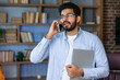 Smiling businessman dressed in blue shirt talking by his phone holding laptop at office. concept of remote and freelance work. Young modern indian successful man in glasses