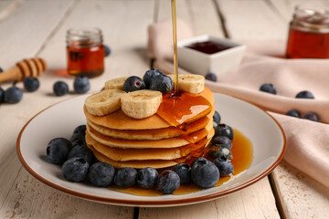Wall Mural - Pouring honey onto tasty pancakes with banana and blueberry on light wooden background, closeup