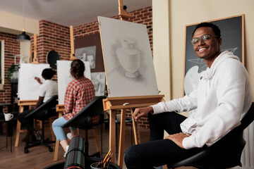 Portrait of happy young African American man student of art school sitting at easel smiling at camera, learning pencil drawing in class. Positive black guy enjoying creative hobby with friends