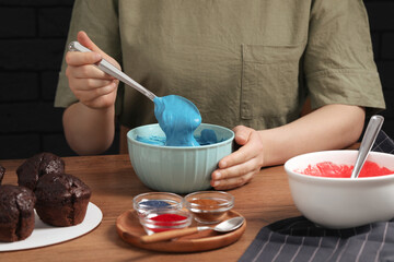 Wall Mural - Woman mixing cream with blue food coloring at wooden table, closeup