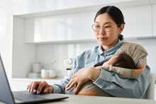 Asian Young Mom Working Online On Laptop While Sitting With Her Baby In Sling In The Kitchen