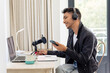Happy biracial transgender man wearing headphones, using laptop, talking to microphone and recording