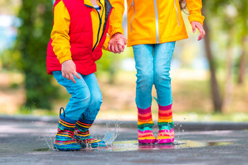 Happy kids girl and boy with umbrella and colorful rubber rain boots playing outdoor and jumping in rainy puddle