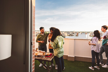 Group of multiracial millennial friends having alcoholic drinks while having outdoor barbecue dinner on the rooftop to meet and celebrate together