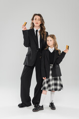 Wall Mural - trendy mother and daughter, businesswoman in suit and schoolgirl in uniform holding credit cards on grey background, modern parenting, financial learning, budgeting, money management