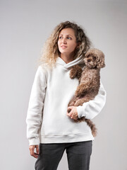 Wall Mural - happy girl with a dog in a photo studio on gray. Curly woman in a white hoodie with a chocolate poodle in her arms.