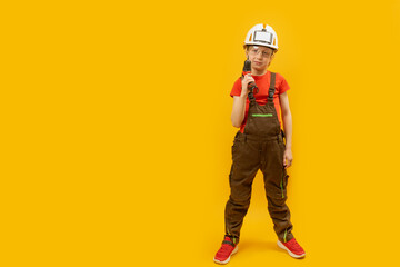 Wall Mural - Child wears protective helmet and holds drill in his hand. Boy as worker on yellow background. Copy space, mock up