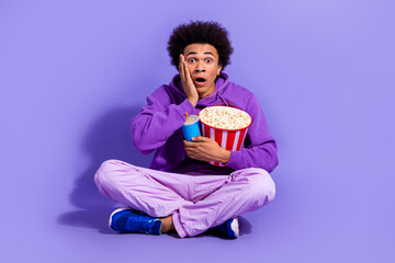 Wall Mural - Full length photo of impressed speechless guy wear purple clothes sit hold popcorn drink arm on cheek isolated on violet color background