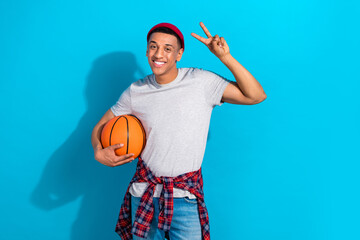 Poster - Photo of cheerful sweet guy dressed grey t-shirt holding ball showing v-sign isolated blue color background