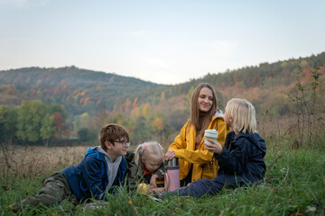 Wall Mural - Happy mom and three kids have fun at picnic in fall. Two brothers with sister and mother drink hot tea on mountains background