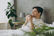 © ViDi Studio - Profile happy young man wear casual clothes t-shirt pajama lying in bed drink coffee tea hold cup close eyes rest relax spend time in bedroom home in own room hotel wake up dream be in good mood day.