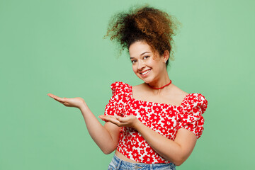 Young smiling happy woman wearing casual clothes red blouse point hands arms aside indicate on workspace area copy space mock up isolated on plain pastel light green color background studio portrait.